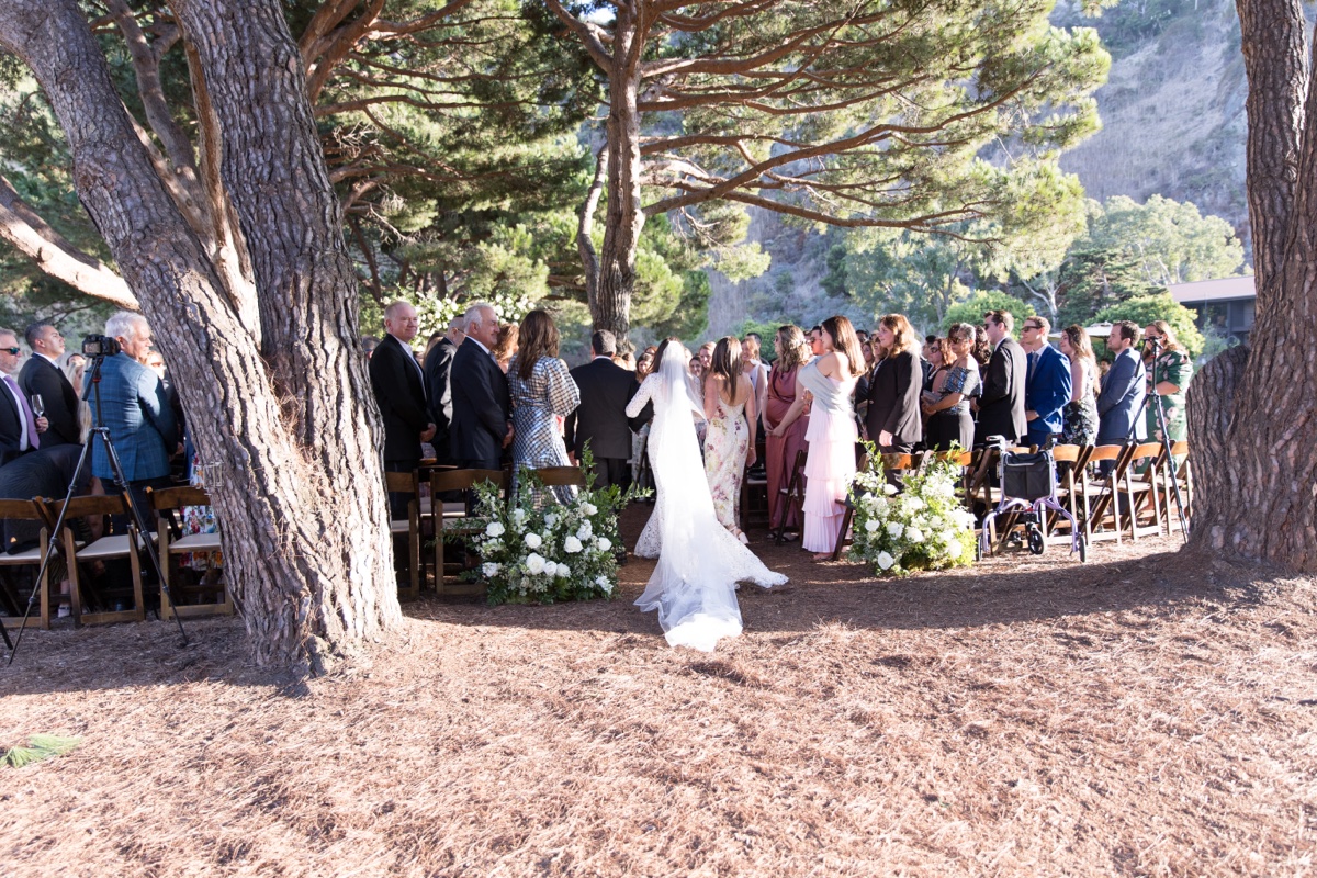 Outdoor wedding ceremony beneath tall trees at Laguna Ranch with white floral arch and elegant details