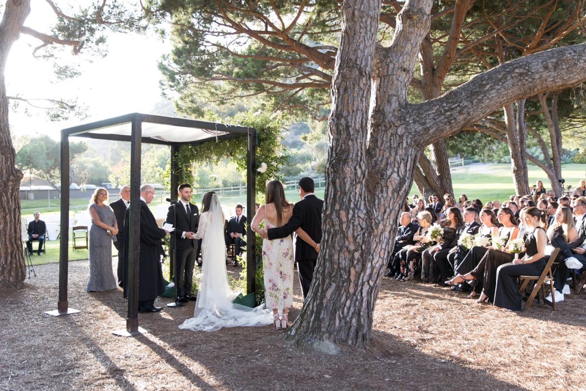 Outdoor wedding ceremony beneath tall trees at ranch at laguna beach with white floral arch and elegant details