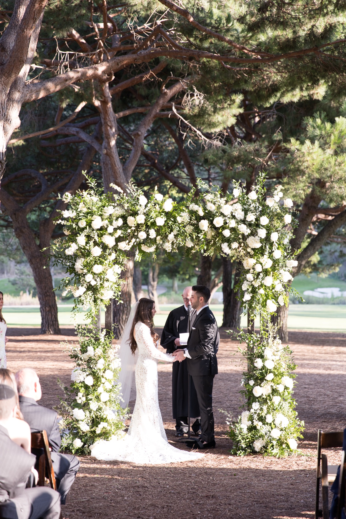 Outdoor wedding ceremony beneath tall trees at ranch at laguna beach with white floral arch and elegant details