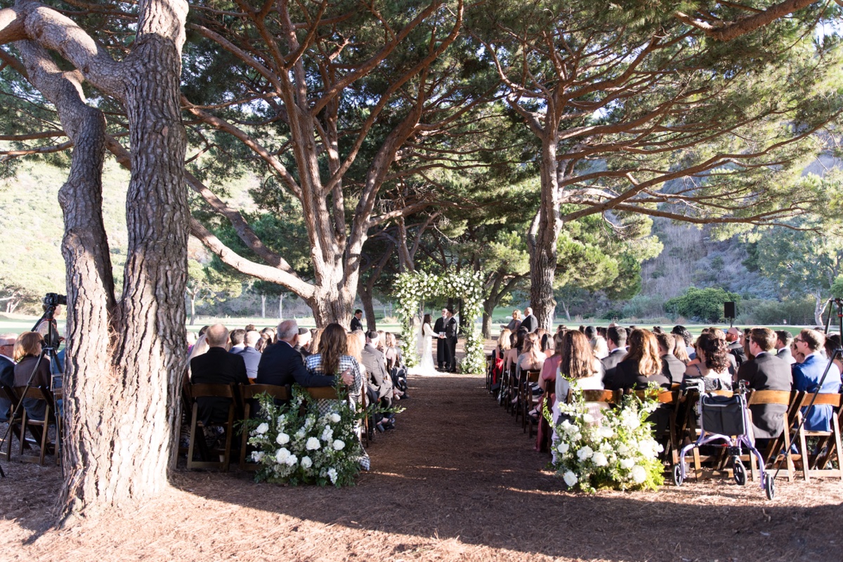 Outdoor wedding ceremony beneath tall trees at ranch at laguna beach with white floral arch and elegant details