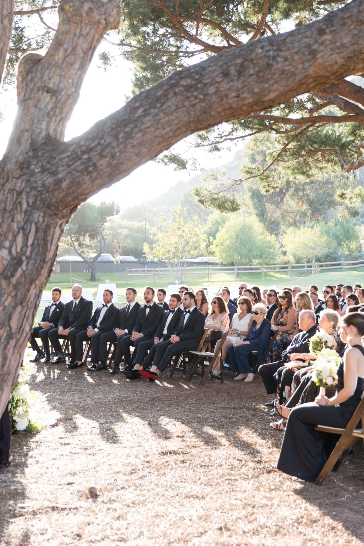 Outdoor wedding ceremony beneath tall trees at ranch at laguna beach with white floral arch and elegant details