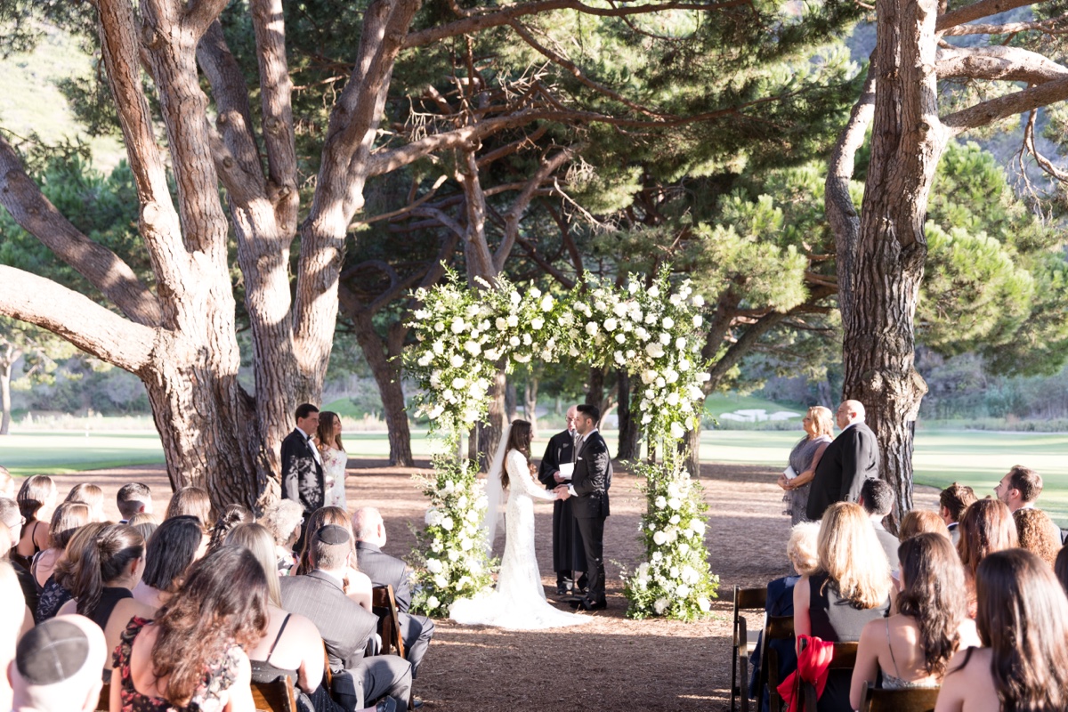 Outdoor wedding ceremony beneath tall trees at ranch at laguna beach with white floral arch and elegant details