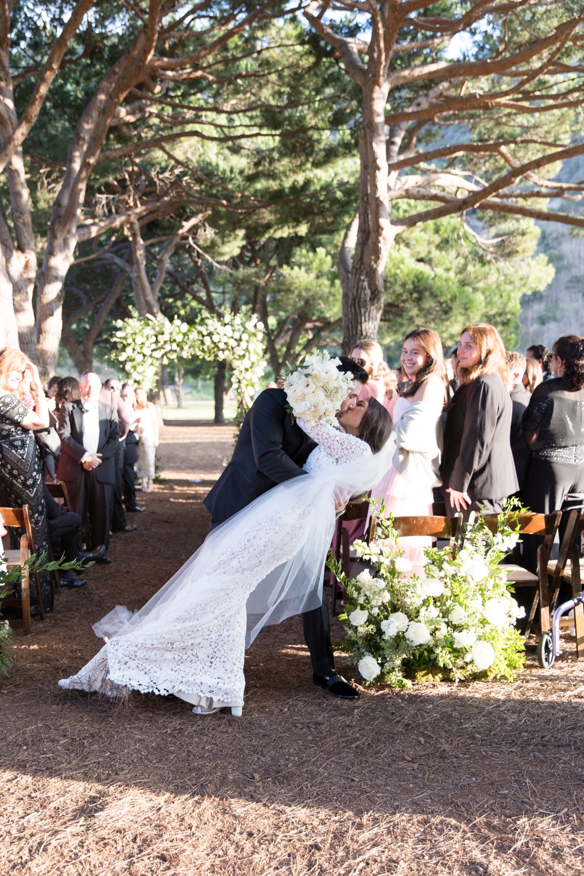 Outdoor wedding ceremony beneath tall trees at ranch at laguna beach with white floral arch and elegant details
